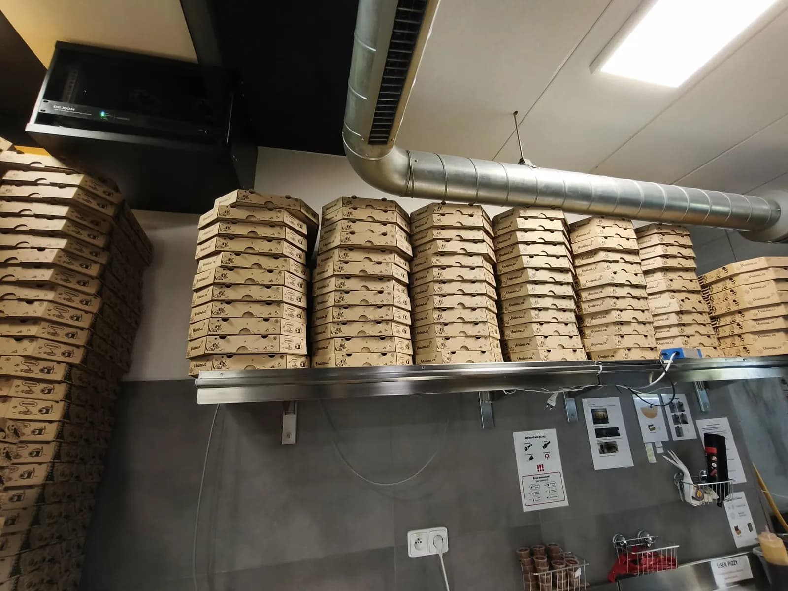 Stacks of pizza boxes on a stainless shelf above the packing station at a Vosíme branch